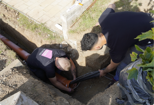 Zwei Heizungstechniker verlegen ein Rohr für eine Wärmepumpe in einem Graben
